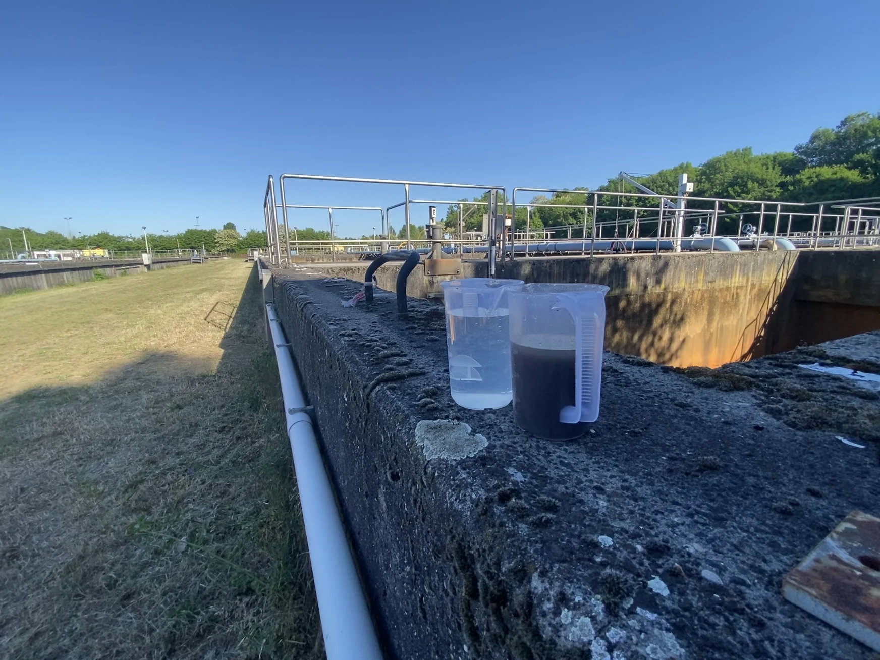 Two water samples on a treatment-basin ledge showing a visible clarity difference, with basin railings, pipework and infrastructure in the background