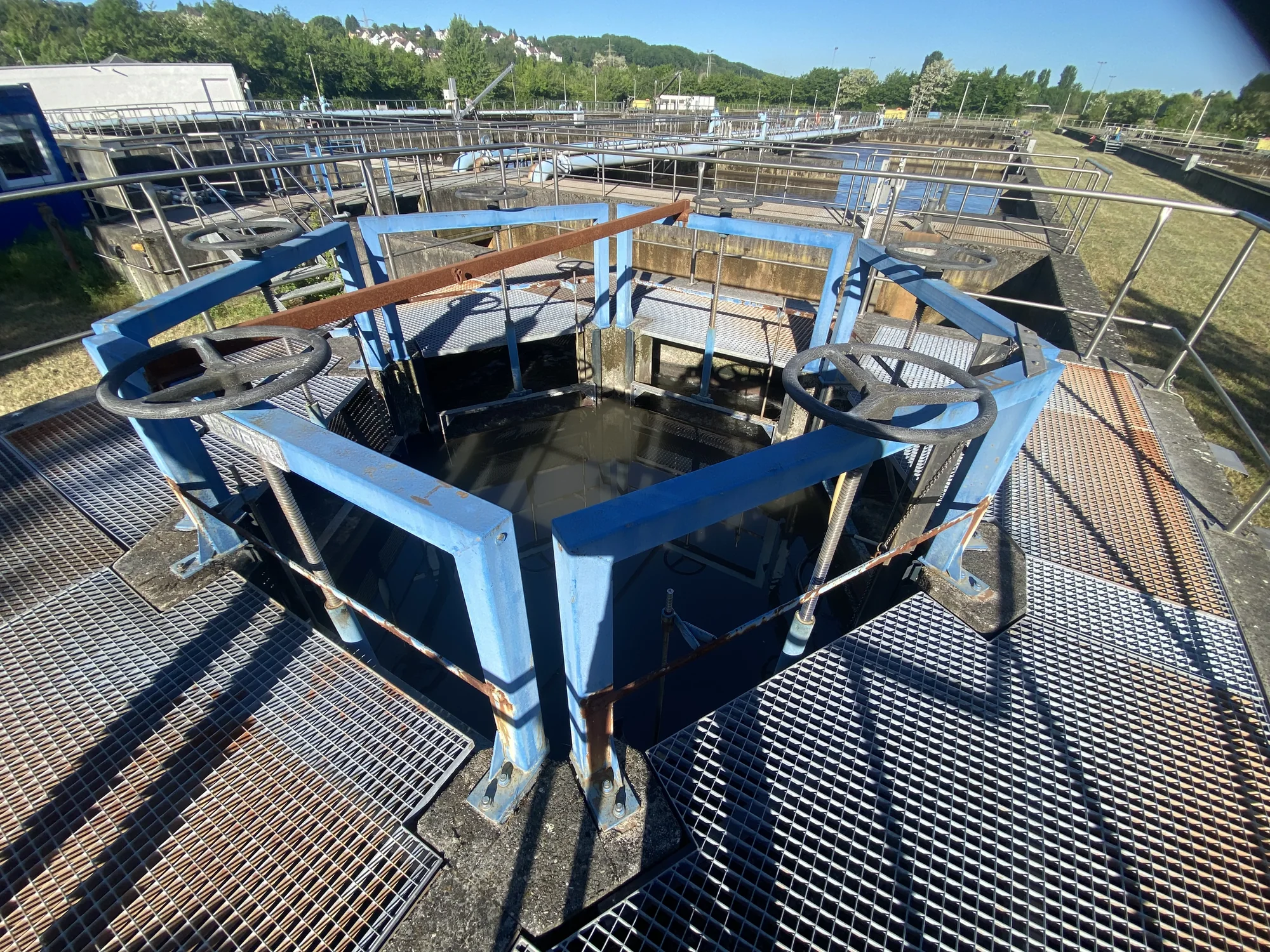 Wide-angle view of a water treatment plant walkway with two manual valve wheels, steel railings and treatment basins in the background